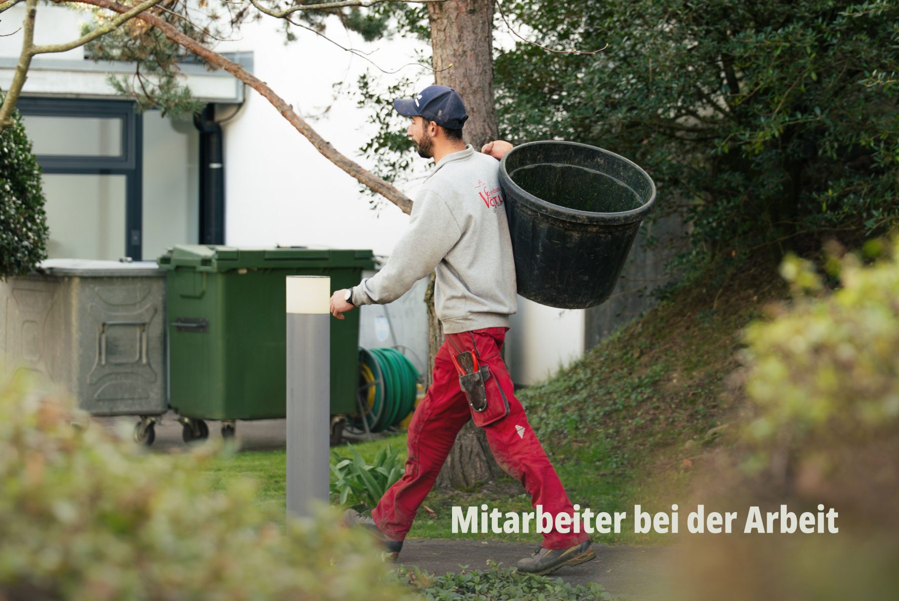 Professionelle Werbefotografie von SR-STUDIOS für Vögtli Gartenbau: Ein Mitarbeiter in roter Arbeitshose trägt einen grossen schwarzen Eimer durch einen gepflegten Garten. Authentische Reportage-Fotografie im hellen Tageslicht. | SR-STUDIOS