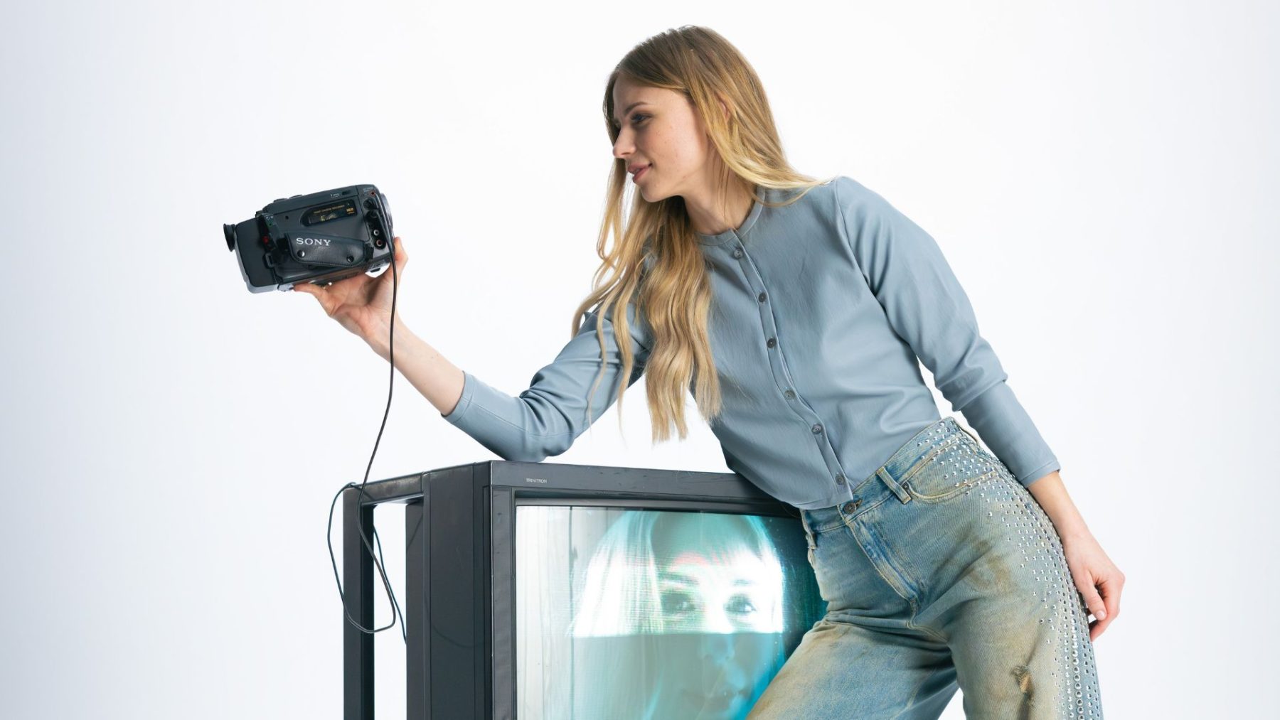Eine junge Frau in einer blauen Strickjacke und Jeans steht neben einem alten Röhrenfernseher, auf dessen Bildschirm ihr eigenes Gesicht spiegelt. Sie hält eine alte Videokamera in der Hand und blickt darauf. Shooting «SR-STUDIOS has great taste». | SR-STUDIOS