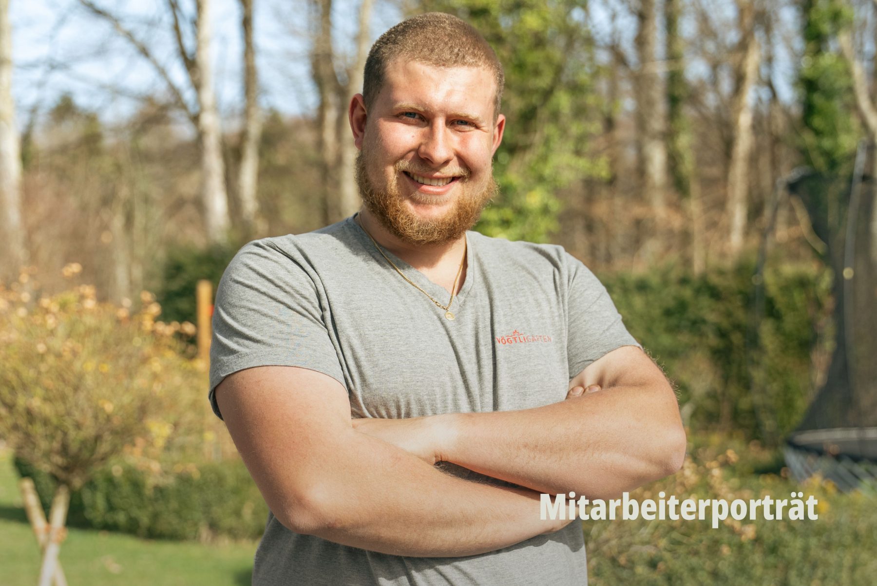Authentisches Mitarbeiterporträt (Employer Branding by SR-STUDIOS): Ein lächelnder, sympatischer Landschaftsgärtner von Vögtli Gartenbau mit verschränkten Armen vor einem natürlichen, grünen Hintergrund im Freien. | SR-STUDIOS