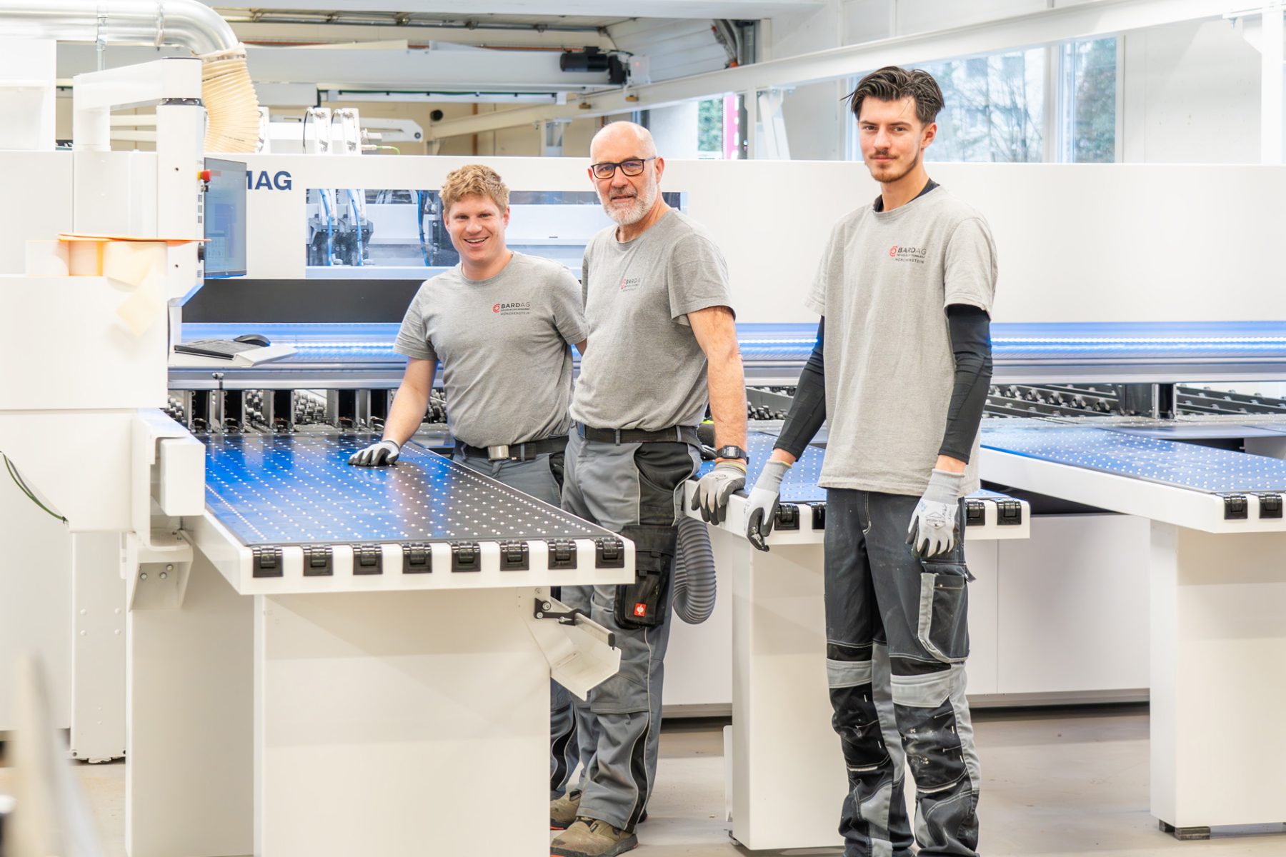 Drei Mitarbeiter, Spezialisten und Schreiner der Bard AG stehen in Arbeitsklamotten in der Werkstatt und bedienen die Zuschnittmaschine. Effiziente Produktion und modernster Maschinenpark für Grossprojekte in der Schweiz. | SR-STUDIOS