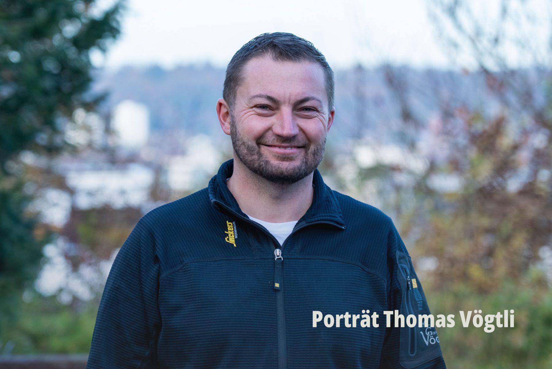 Portraitfotografie für Unternehmen: Thomas Vögtli steht freundlich lächelnd in einer dunklen Arbeitsjacke vor einem weichgezeichneten, natürlichen Hintergrund. Employer Branding by SR-STUDIOS. | SR-STUDIOS