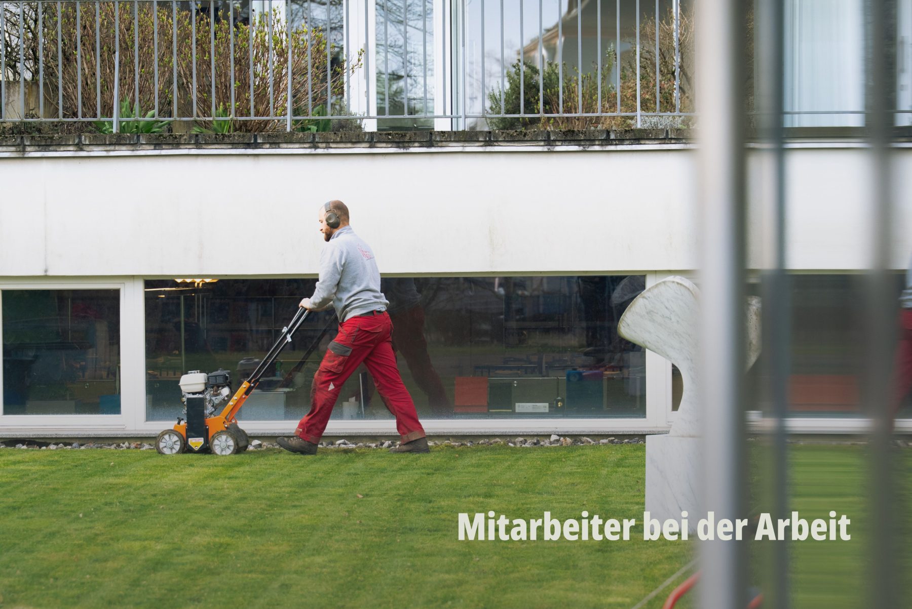 Reportagefoto aus dem Arbeitsalltag: Ein Gärtner der Vögtli Gartenbau GmbH mäht den Rasen vor einem modernen Gebäude. Fotografiert von SR-STUDIOS durch ein architektonisches Element im Vordergrund für zusätzliche Bildtiefe. | SR-STUDIOS